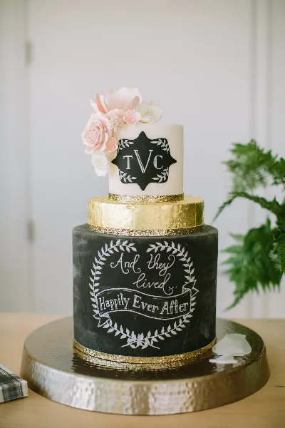 A two-tier wedding cake with a black bottom tier reading 'And they lived Happily Ever After' and a white top tier with pink sugar flowers and the initials TVC.