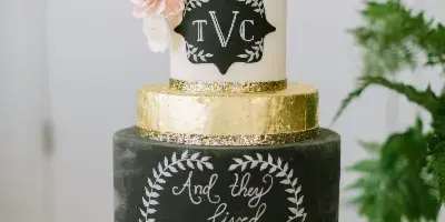 A two-tier wedding cake with a black bottom tier reading 'And they lived Happily Ever After' and a white top tier with pink sugar flowers and the initials TVC.