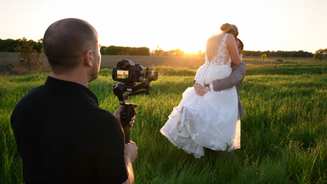A videographer filming a bride and groom in a field at sunset