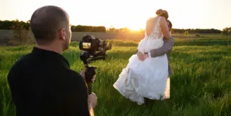 A videographer filming a bride and groom in a field at sunset