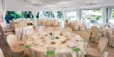 A large white marquee tent interior set up for a formal event with round tables, white tablecloths, chandeliers, and place settings.