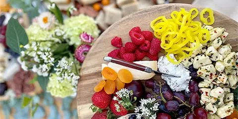A close-up of a catering platter featuring fresh raspberries, strawberries, grapes, cheese, and crackers, garnished with small white flowers.