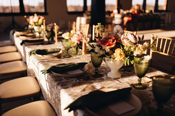 Styled wedding reception table with patterned linens, green glassware, candles, place settings, and floral centerpieces.