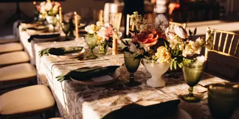 Styled wedding reception table with patterned linens, green glassware, candles, place settings, and floral centerpieces.