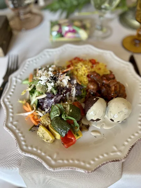 A plated catering dish featuring salad, rice, roasted vegetables, and a meat entrée on a white plate at a set table.