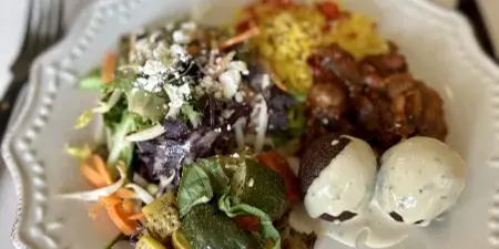 A plated catering dish featuring salad, rice, roasted vegetables, and a meat entrée on a white plate at a set table.