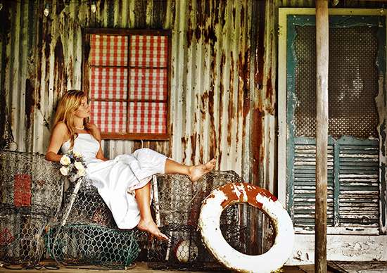 A staged event set featuring a bride in a white dress seated on a wire-mesh sofa against a weathered, rustic wall with a window and a life preserver.