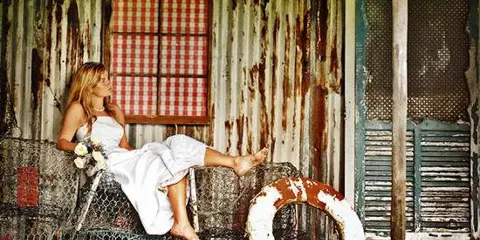 A staged event set featuring a bride in a white dress seated on a wire-mesh sofa against a weathered, rustic wall with a window and a life preserver.