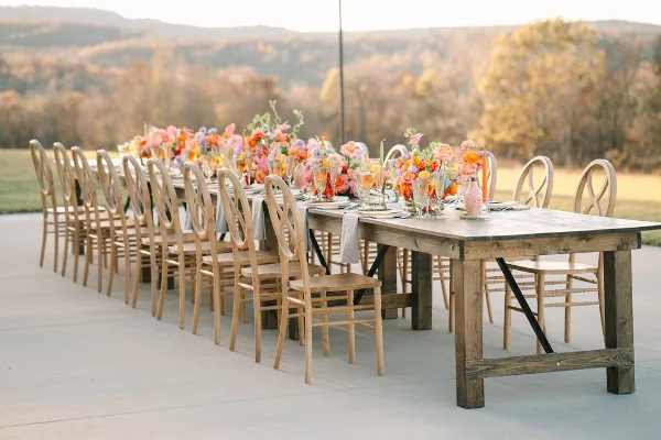A long wooden dining table set with floral arrangements and chairs on an outdoor patio overlooking a scenic landscape of hills and trees.