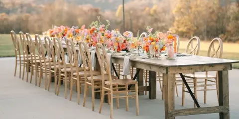 A long wooden dining table set with floral arrangements and chairs on an outdoor patio overlooking a scenic landscape of hills and trees.