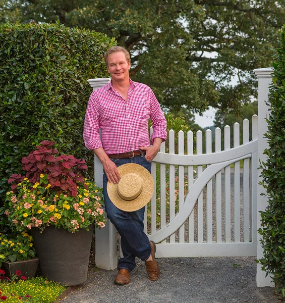 P. Allen Smith standing at the white picket gate of Moss Mountain Farm with a straw hat and potted flowers.