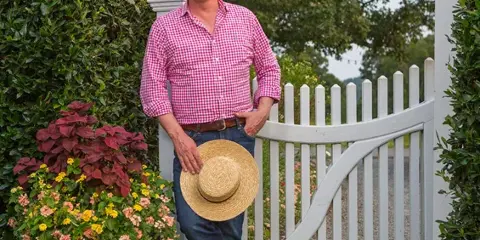 P. Allen Smith standing at the white picket gate of Moss Mountain Farm with a straw hat and potted flowers.