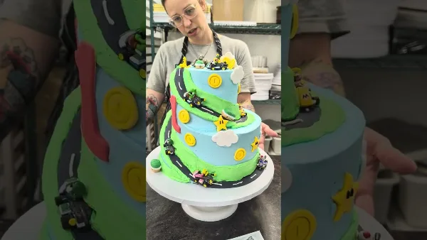 A baker in a kitchen holding a multi-tiered custom cake decorated with a Mario Kart theme, featuring fondant roads, coins, and characters.