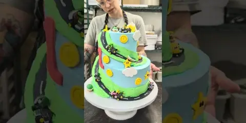 A baker in a kitchen holding a multi-tiered custom cake decorated with a Mario Kart theme, featuring fondant roads, coins, and characters.