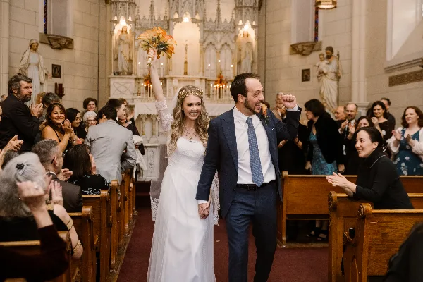 A bride and groom walking down the aisle of a church during a wedding ceremony while guests applaud.