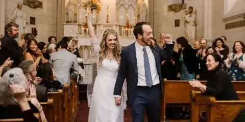 A bride and groom walking down the aisle of a church during a wedding ceremony while guests applaud.