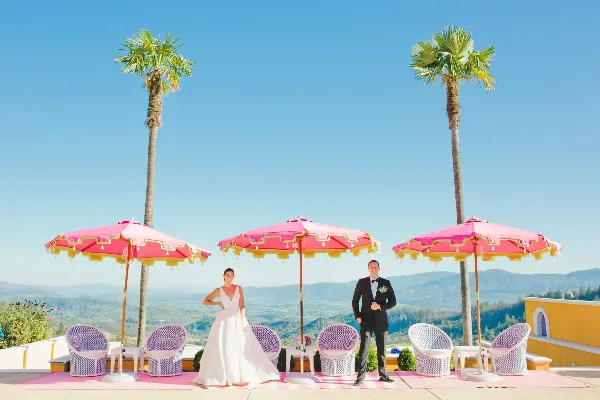 A bride and groom standing on a terrace with pink umbrellas and a mountain view.