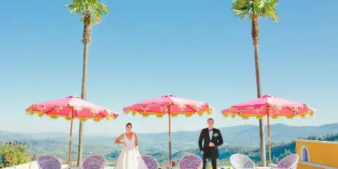 A bride and groom standing on a terrace with pink umbrellas and a mountain view.