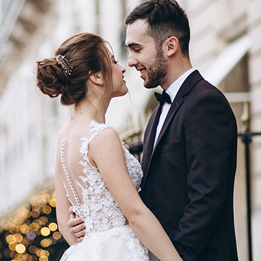 A bride and groom smiling at each other in a romantic setting with blurred lights in the background.