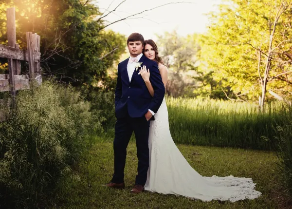 A bride and groom posing together in a grassy field with a wooden fence in the background.