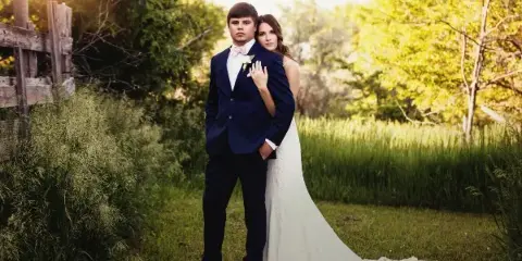 A bride and groom posing together in a grassy field with a wooden fence in the background.