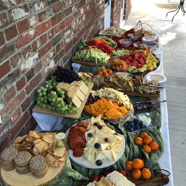 A long table set with a diverse catering spread including a cheese and fruit platter, charcuterie, crackers, and fresh vegetables.
