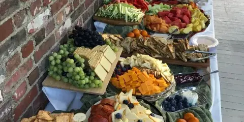 A long table set with a diverse catering spread including a cheese and fruit platter, charcuterie, crackers, and fresh vegetables.