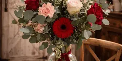 A floral arrangement of red and white roses, a red gerbera daisy, and eucalyptus in a glass vase tied with a red ribbon, displayed on a wooden table.