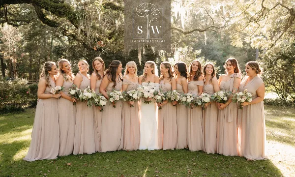 A bride and bridesmaids in beige dresses holding white and green floral bouquets in a wooded outdoor setting.