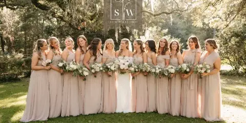 A bride and bridesmaids in beige dresses holding white and green floral bouquets in a wooded outdoor setting.
