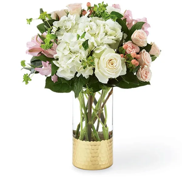 A floral arrangement featuring white hydrangeas, pink and white roses, and greenery in a clear glass vase with a gold base.