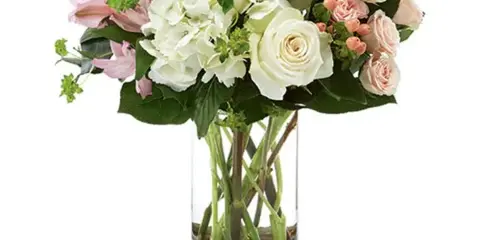 A floral arrangement featuring white hydrangeas, pink and white roses, and greenery in a clear glass vase with a gold base.