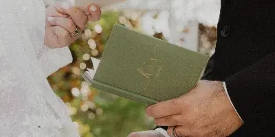 A close-up photograph of a bride and groom exchanging rings and holding vow books during an outdoor wedding ceremony.