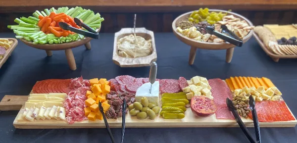 A catering table display featuring a large charcuterie board with meats, cheeses, olives, and pickles, alongside bowls of fresh vegetables and dips.