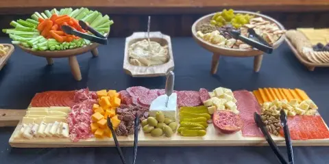 A catering table display featuring a large charcuterie board with meats, cheeses, olives, and pickles, alongside bowls of fresh vegetables and dips.