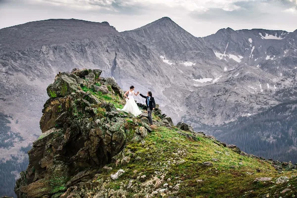 A bride and groom standing on a rocky mountain peak with a vast mountain range in the background.