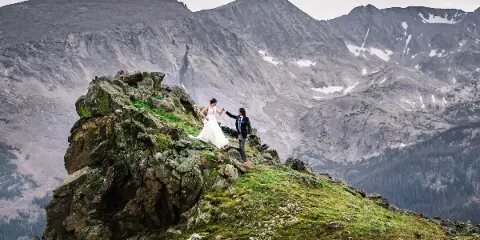 A bride and groom standing on a rocky mountain peak with a vast mountain range in the background.