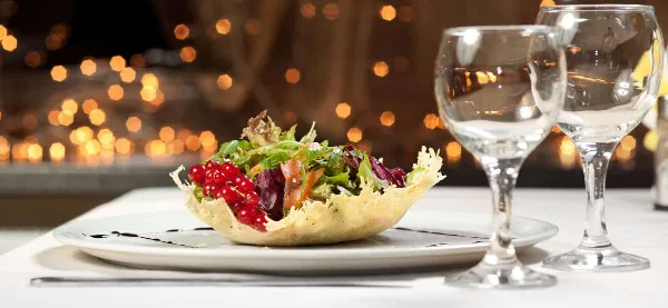 A plated salad in a pastry bowl with red currants and greens, served with two wine glasses on a white tablecloth.