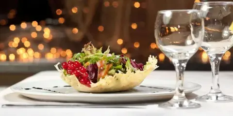 A plated salad in a pastry bowl with red currants and greens, served with two wine glasses on a white tablecloth.