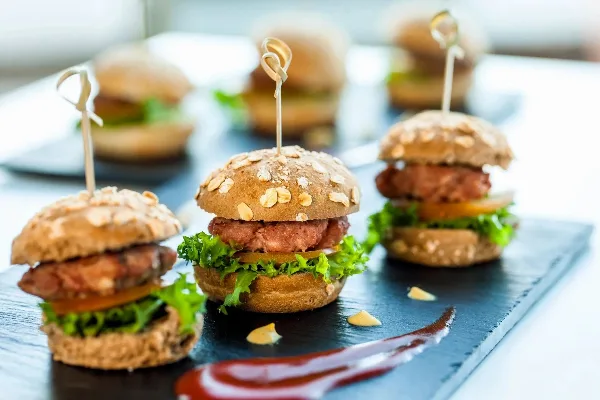 A close-up photograph of several mini sliders with oat-topped buns, lettuce, and meat patties arranged on a dark slate serving board.