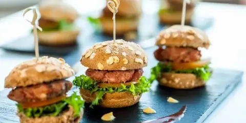 A close-up photograph of several mini sliders with oat-topped buns, lettuce, and meat patties arranged on a dark slate serving board.