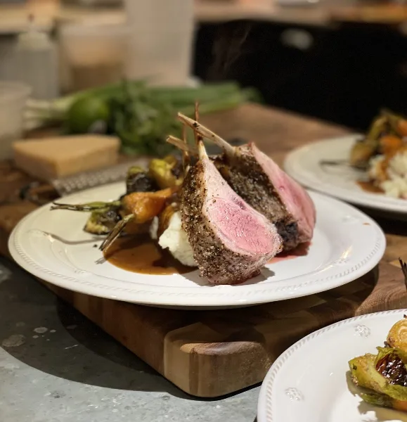 A plated dish featuring a seared lamb chop with a peppercorn crust, served with roasted vegetables and mashed potatoes on a white plate.