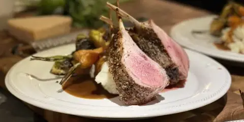 A plated dish featuring a seared lamb chop with a peppercorn crust, served with roasted vegetables and mashed potatoes on a white plate.