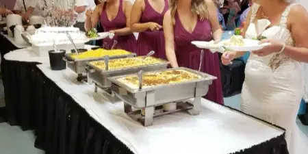 A bride and bridesmaids serving food from a buffet line at a wedding reception.