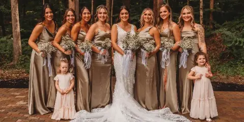 A bride and bridesmaids in champagne dresses posing in a forest setting with a wooden structure.