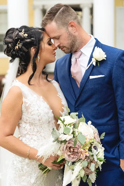 A bride in a lace gown and a groom in a blue suit standing close together, holding a bouquet of white and pink flowers.