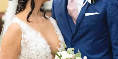 A bride in a lace gown and a groom in a blue suit standing close together, holding a bouquet of white and pink flowers.