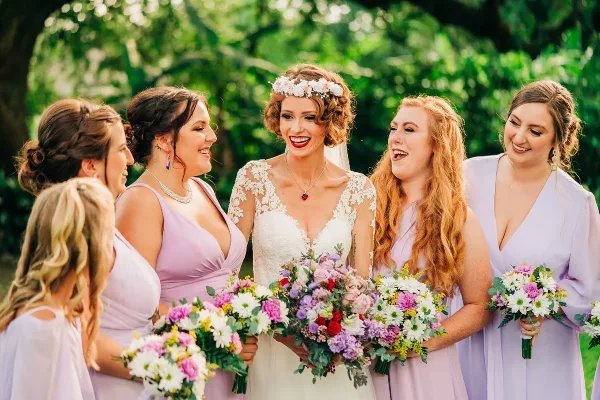 A bride in a white lace gown and floral crown laughing with her bridesmaids in lavender dresses, holding colorful bouquets outdoors.