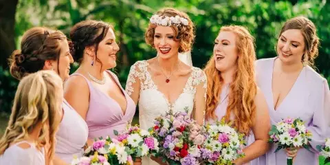 A bride in a white lace gown and floral crown laughing with her bridesmaids in lavender dresses, holding colorful bouquets outdoors.