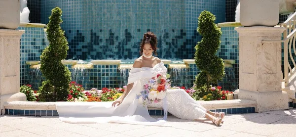 Bride in a white gown seated by a fountain holding a colorful bridal bouquet